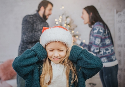 Child wearing a Santa hat covers their ears while parents argue in the background, illustrating the impact of holiday custody disputes on children.