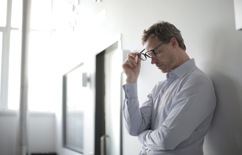 tired looking businessman leaning against wall taking off glasses